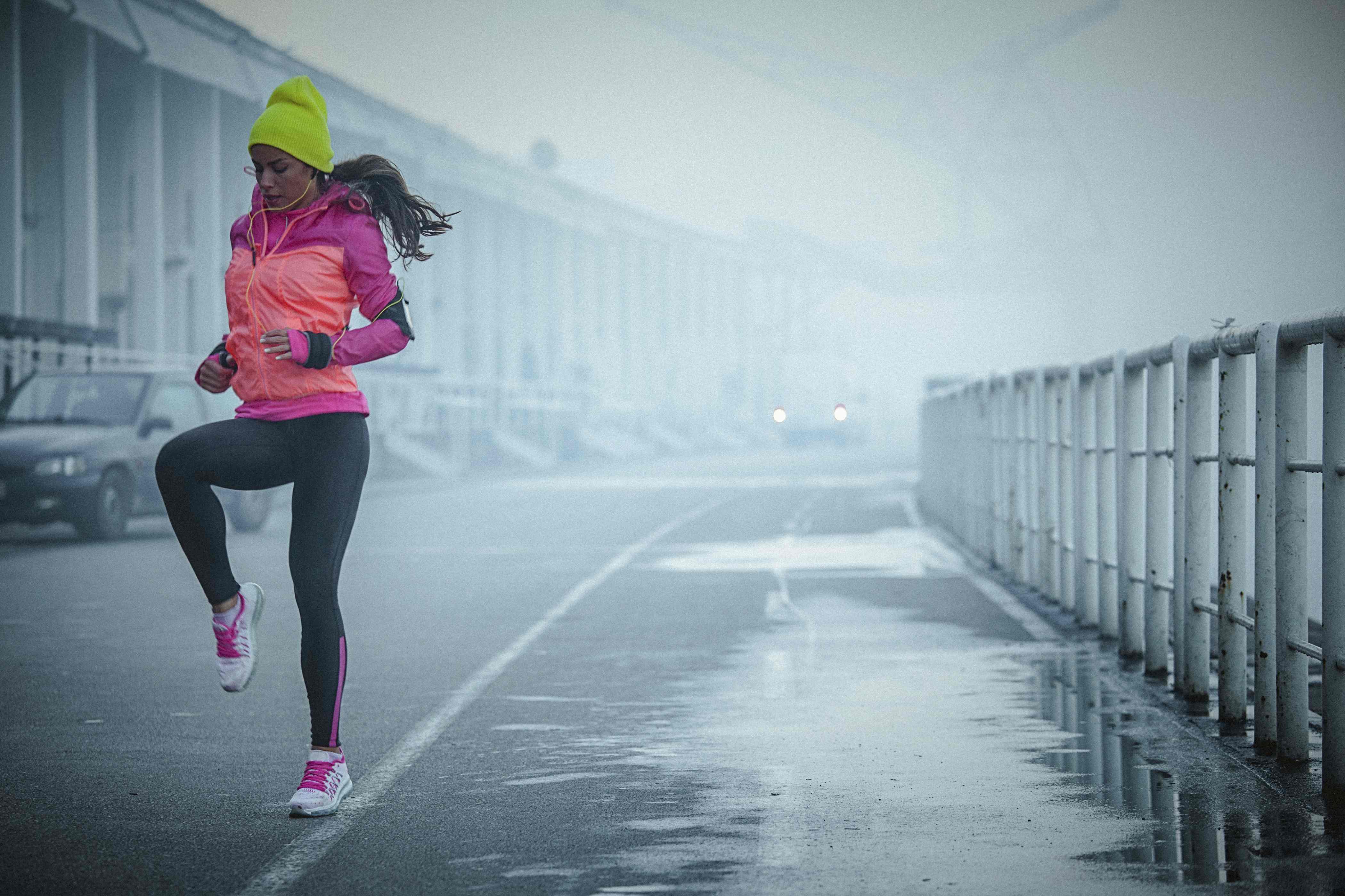 Young athlete woman exercises bouncing up and down and listens to music in the city port. The woman wears black running tights and red jacket. She has a smart phone mounted on her arm, with earphones connected to it. Her body is in motion, slightly above the ground. The copy space has been left. Shallow DOF.