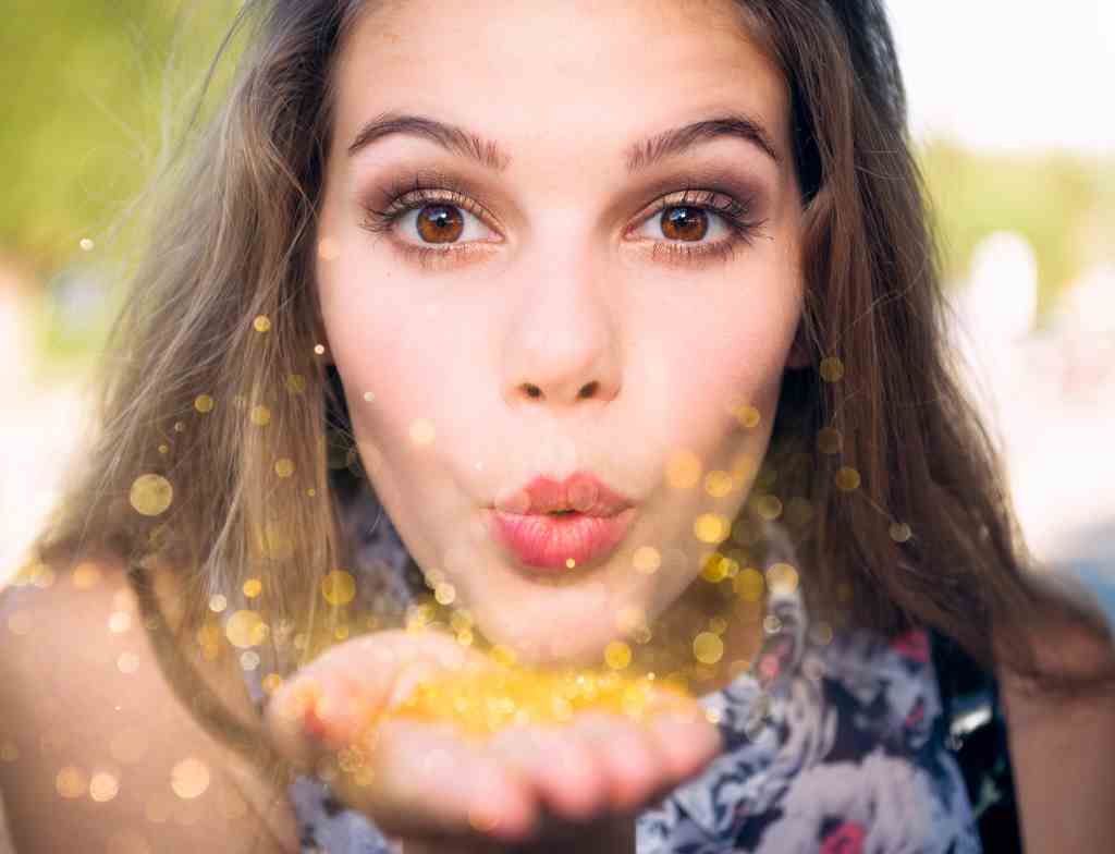 PLESES MRMRM A young woman blowing gold glitter from her hand, towards the camera.